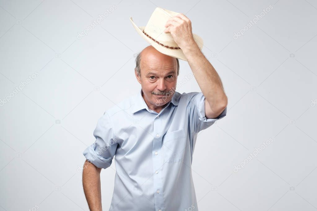 You are welcome concept. Cheerful mature man in blue shirt and cowboy hat gesturing welcome sign and smiling while standing against gray wall. He is happy to meet dear guests