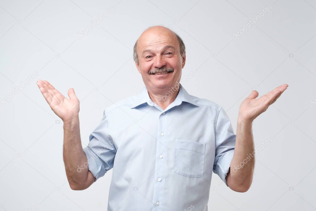 Indoor portrait of confused senior man in blue shirt showing I have no idea gesture, shrugging shoulders and raising hands, standing against gray background. Sorry I did not mean it