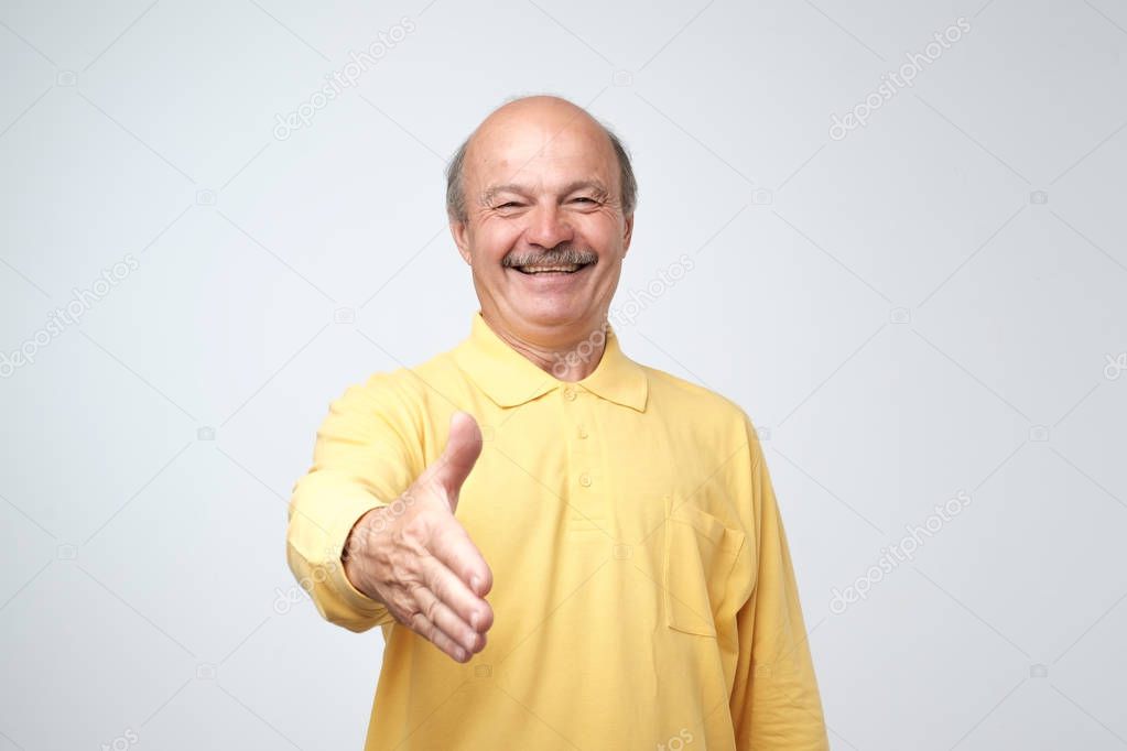 You are welcome concept. Cheerful mature man in yellow t-shirt shirt gesturing welcome sign and smiling while standing against gray wall. He is happy to meet dear guests