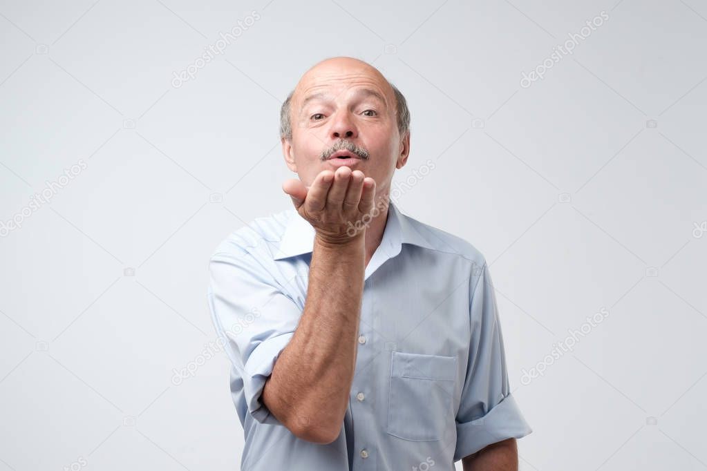 Bald handsome mature male in blue shirt blowing air kiss, expresses his devotion and truthful love to his wife isolated over white background. People, body language and facial expressions concept