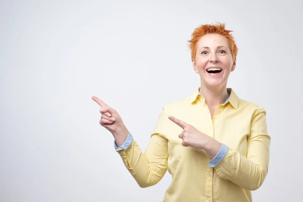 Surprised funny mature woman in yellow shirt showing empty copy space with her finger grey background. — Stock Photo, Image