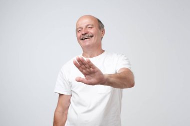 Portrait of laughing mature man wearing white t-shirt showing stop gesture with palm, laughing on funny joke. Please stop amuse me. People sincere emotions lifestyle concept. clipart