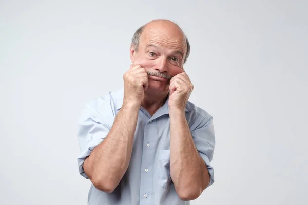 Senior spansktalande mannen i blå skjorta dra huden på ansikte med händerna. Han är rädd eller trött av stress. — Stockfoto