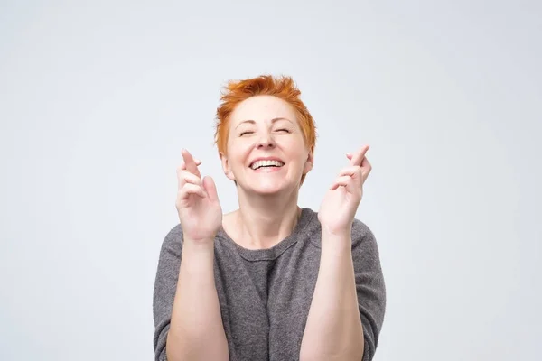 Kobieta szczęśliwa dojrzałe rude chętny do wygrania konkurencji, trzyma kciuki, jako czeka na wyniki i uśmiecha się szeroko — Zdjęcie stockowe