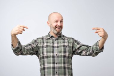 Yüzünde gülümseme ile kendine güvenen seyir gri grunge duvarın üzerinden sakal ayakta olgun kel yetişkin adam. O biraz bencil ve kendini, parmaklarıyla işaret ediyor