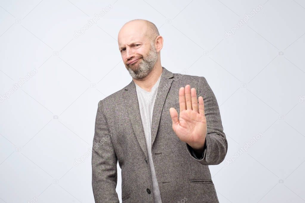 Attractive mature man showing refusal gesture. It is not for me, leave me in piece, has angry expression, poses against white concrete studio background