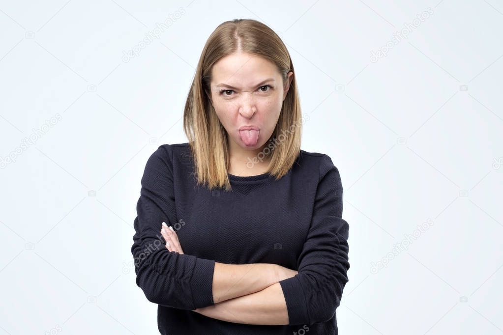 Dissatisfied woman frowns face, has disgusting expression, shows tongue, expresses disgust, irritated with somebody