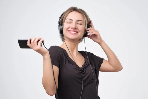 Preciosa mujer escuchando música en auriculares y cantando sobre fondo gris . — Foto de Stock