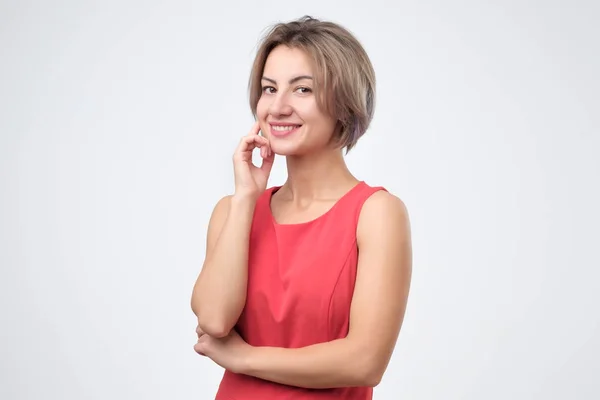Mujer atractiva carismática en vestido rojo, sonriendo alegremente mientras está de pie sobre fondo gris —  Fotos de Stock
