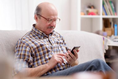 Olgun Latin asıllı adam cep telefonuyla ev oturma odasında kanepede oturan mesajlarını gözlük takıyor