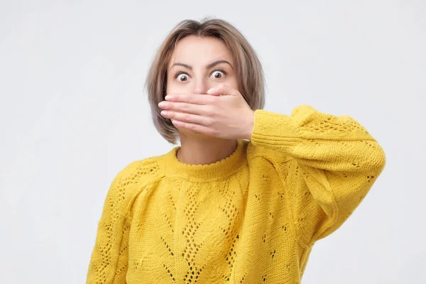 Verrast vrouw in gele trui die betrekking hebben op mond met de hand en camera staren — Stockfoto
