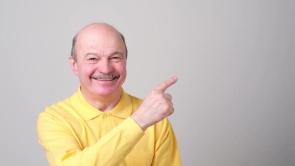 Uomo anziano guardando la macchina fotografica, sorridendo e indicando da parte con mano . — Video Stock