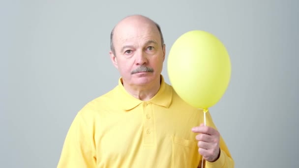 A sad senior man in a yellow shirt is holding a small balloon in his hands. — Stock Video