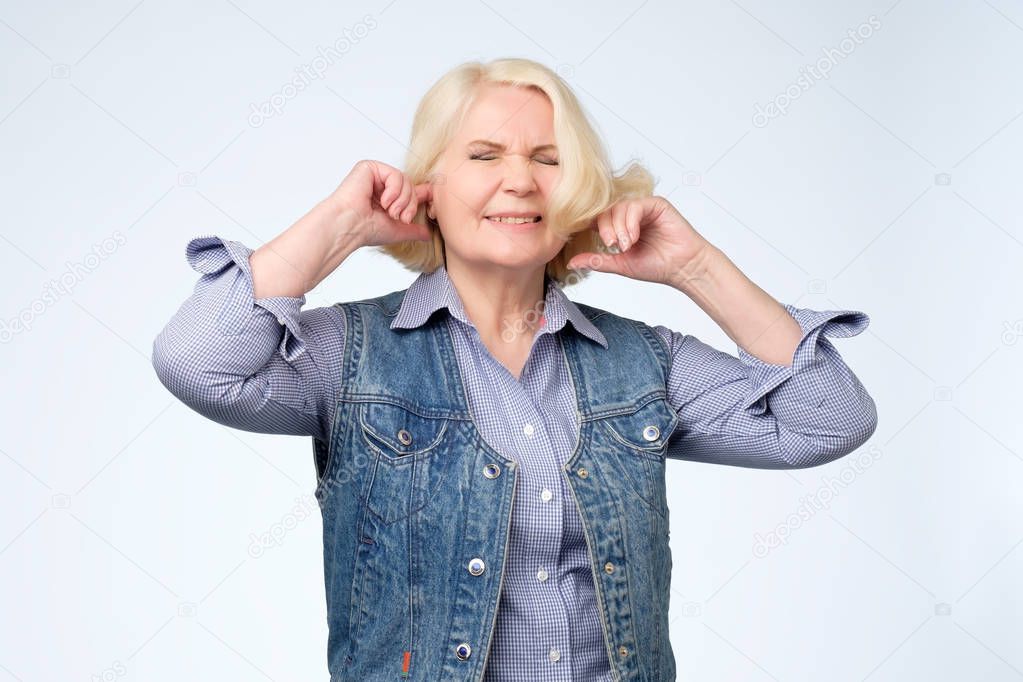 unhappy stressed out senior female making wry face, plugging ears with fingers