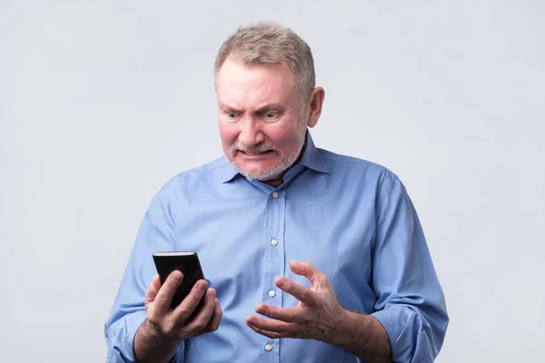 Annoyed senior man in blue shirt looking at mobile phone — Stock Photo, Image