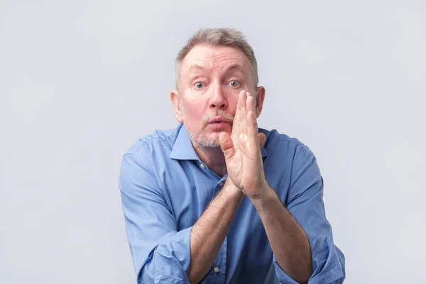 Hombre mayor con camisa azul cogido de la mano en la boca contando rumores secretos —  Fotos de Stock