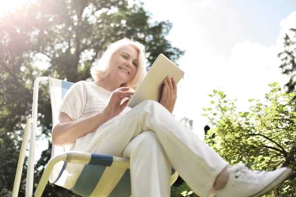 Senior blonde vrouw zittend in de stoel en het gebruik van digitale Tablet in de tuin — Stockfoto