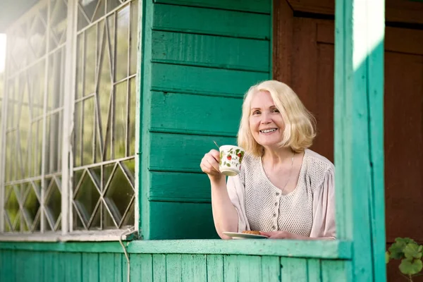 Старша жінка відпочиває і п'є чай, сидячи на терасі літнього будинку — стокове фото