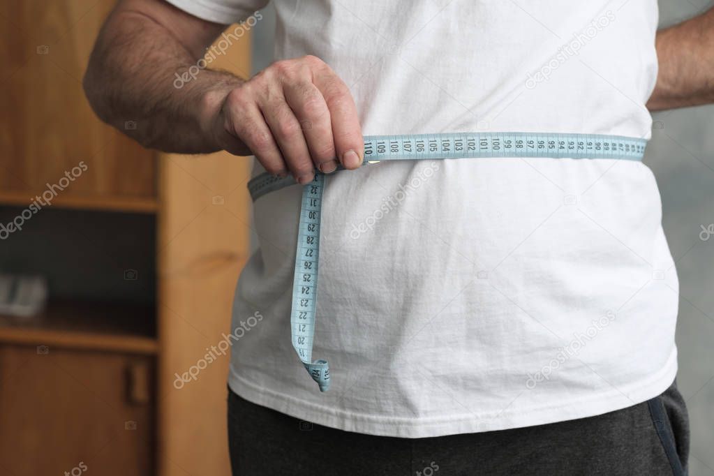 Man measuring his belly with measurement tape standing in the living room.