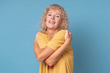 Rahatlamış olgun bir kadın, neşeli bir yüz ifadesi takınarak kendine sarılıyor.