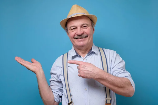 Senior sorrindo feliz homem mostra algo no fundo azul . — Fotografia de Stock