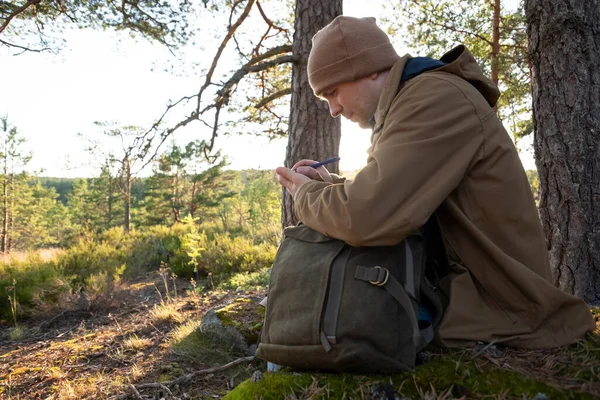 man in forest sits on the ground, writing something in diary.