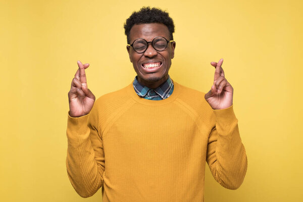 Young african man making wish gesture holding fingers crossed. Studio shot on yellow wall.