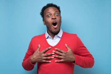 Dost canlısı Afrikalı Amerikalı adam ellerini kalbinin üstünde tutuyor, iltifatlarla dokunuyor, minnettarlığını gösteriyor, ifadesini memnun ediyor. Stüdyo resmi mavi duvarda.