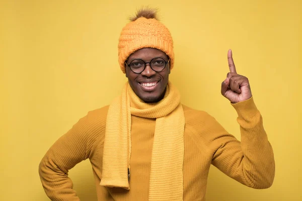 Satisfied african man in swinter clothes showing index fingers up, giving advice or recommendation. Studio shot on yellow wall.