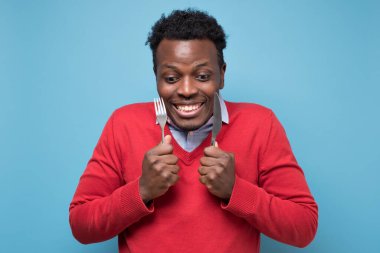 Çatal ve bıçak tutan aç Afrikalı bir adam yemek için bekliyor. Stüdyo resmi mavi duvarda.