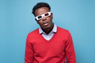 3 boyutlu gözlüklü Afrikalı genç adam daha kalın kafalıların sersemlemesini izliyor. Stüdyo resmi mavi duvarda.