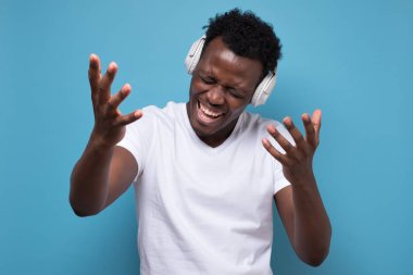 Beyaz kulaklıklarla müzik dinleyen yakışıklı Afro-Amerikan bir genç. Müzik konseptinin tadını çıkar Stüdyo resmi mavi duvarda.