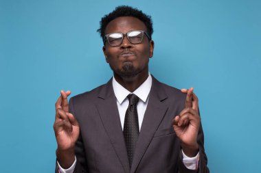 Afrikalı genç işadamı mavi arka planda parmaklarını çapraz yapıyor.