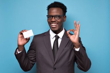 Genç gülümseyen Afrikalı adam kredi kartı tutuyor ve mavi arka planda iyi işareti gösteriyor.