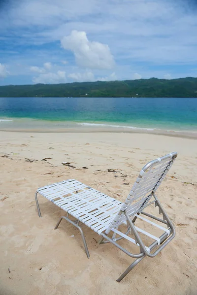 Small plastic deck chair standing on sand of Cayo Levantado beach against beautiful water of Atlantic Ocean in the Samana Bay. Lounge is white and lightwaight. water is blue and sky has some clouds.