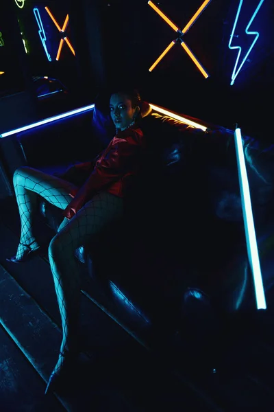 Cyberpunk shooting di modella vestita di giacca rossa da motociclista seduta in divano di pelle contro neon — Foto Stock