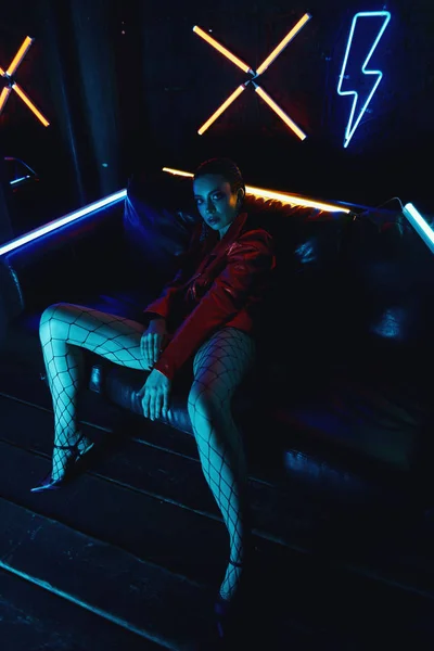 Cyberpunk skytte av modell klädd i röd cyklister jacka sitter i skinnsoffa mot Neon — Stockfoto