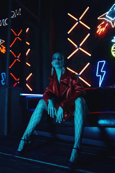 Kyberpunkové natáčení modelu s červeným bikerovým kabátem sedícího v kožené pohovce proti neonům — Stock fotografie