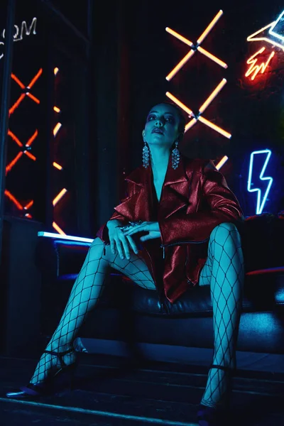 Cyberpunk skytte av modell klädd i röd cyklister jacka sitter i skinnsoffa mot Neon — Stockfoto