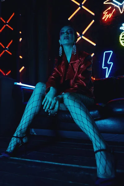 Cyberpunk shooting of model wearing red bikers jacket sitting in leather sofa against neon — Stock Photo, Image