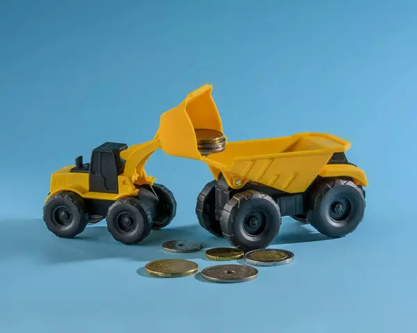 Yellow toy bulldozer loading coins money to yellow toy truck. Space for text. Blue background