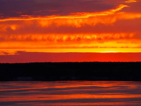 A fiery sunset is reflected in the calm river water, overhanging clouds in the bright orange sky, light waves on the water surface.