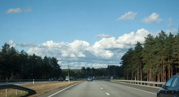 view on asphalt road with cars and big clouds in blue sky and forest trees around