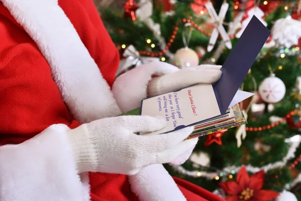 closeup of santa claus holding stack of letters and postcards from children and reading them in front of christmas tree