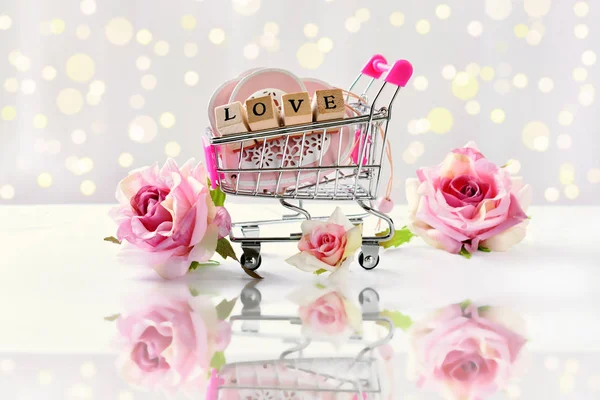shopping cart filled with hearts and wooden cubes with letters LOVE and roses on white background with bokeh