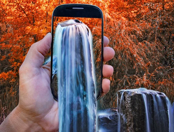 Digitales Zusammenspiel einer Hand mit Smartphone und Wasserfall im Herbst — Stockfoto