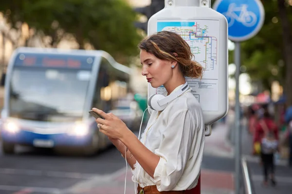 城市公共汽车站配备音乐耳机电话应用程序的妇女 — 图库照片