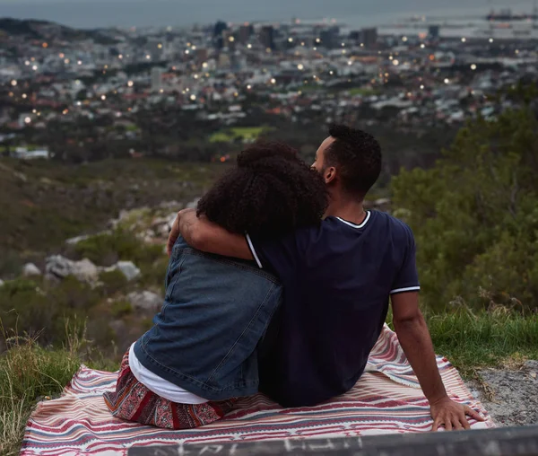 back view of couple looking at city from hill and sitting on blanket, man hugging woman