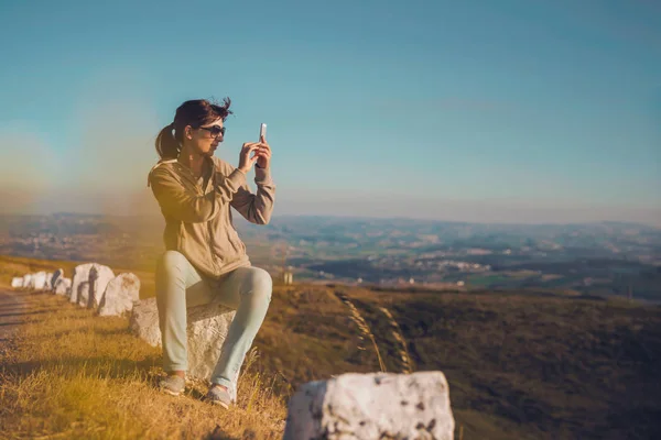 美丽的女人拍照与智能手机 而坐在石头上在山上 — 图库照片