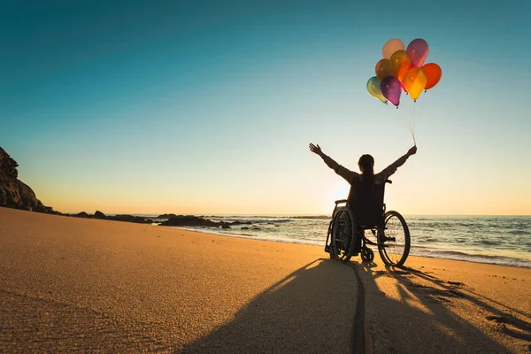 坐在轮椅上 沙滩上拿着彩色气球的残疾妇女 — 图库照片